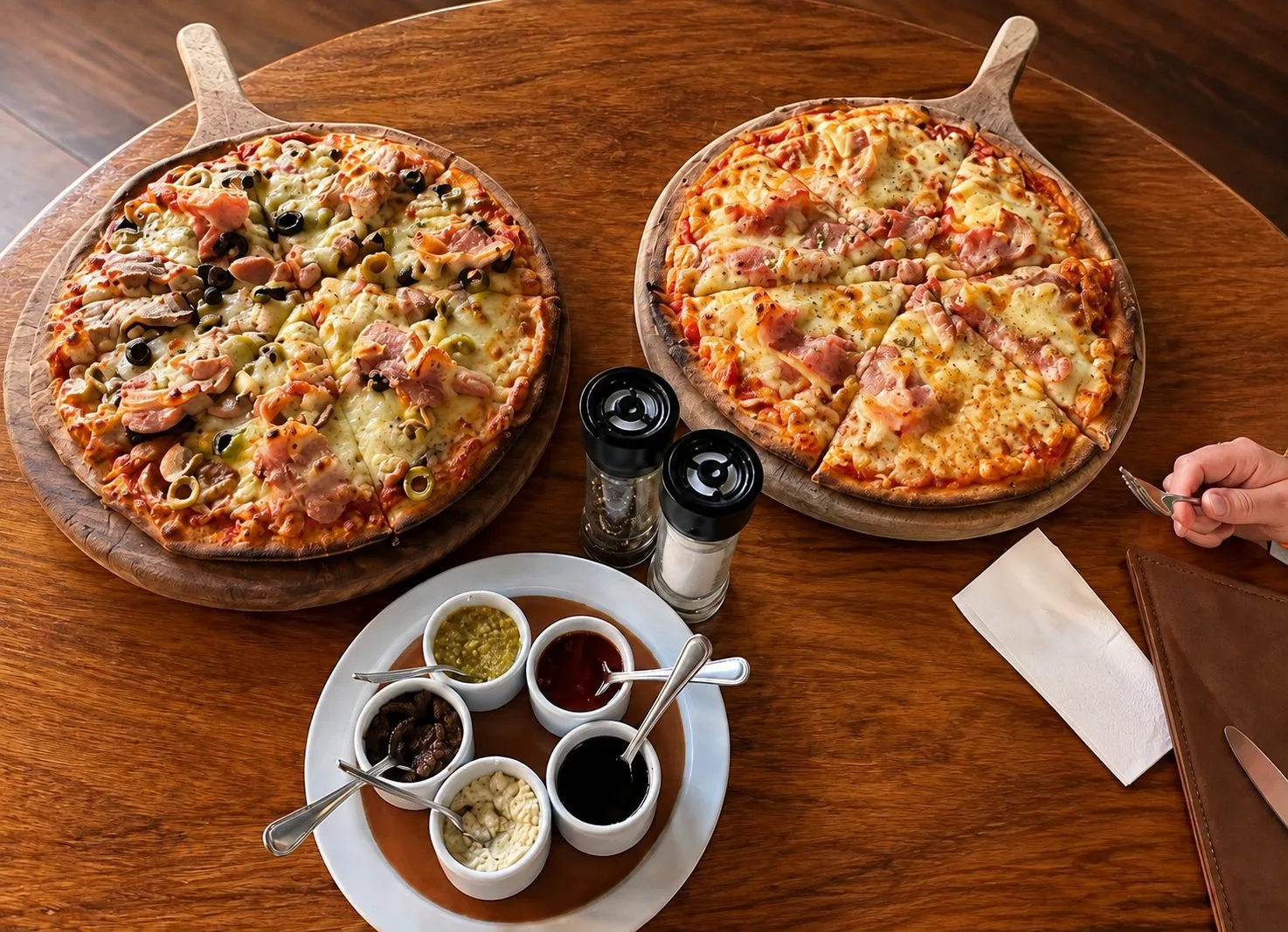 Dos pizzas recién horneadas sobre tablas de madera, con la tabla de salsas de la casa